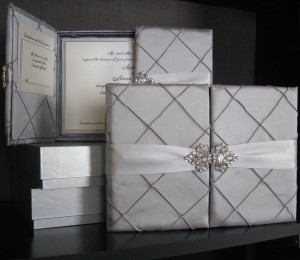 Padded silver pintuck fabric box has a double rhinestone bling closure, with pockets to hold insert cards and a 3-layer pearl/silver invitation floating on a hidden 3-dimensional riser inside